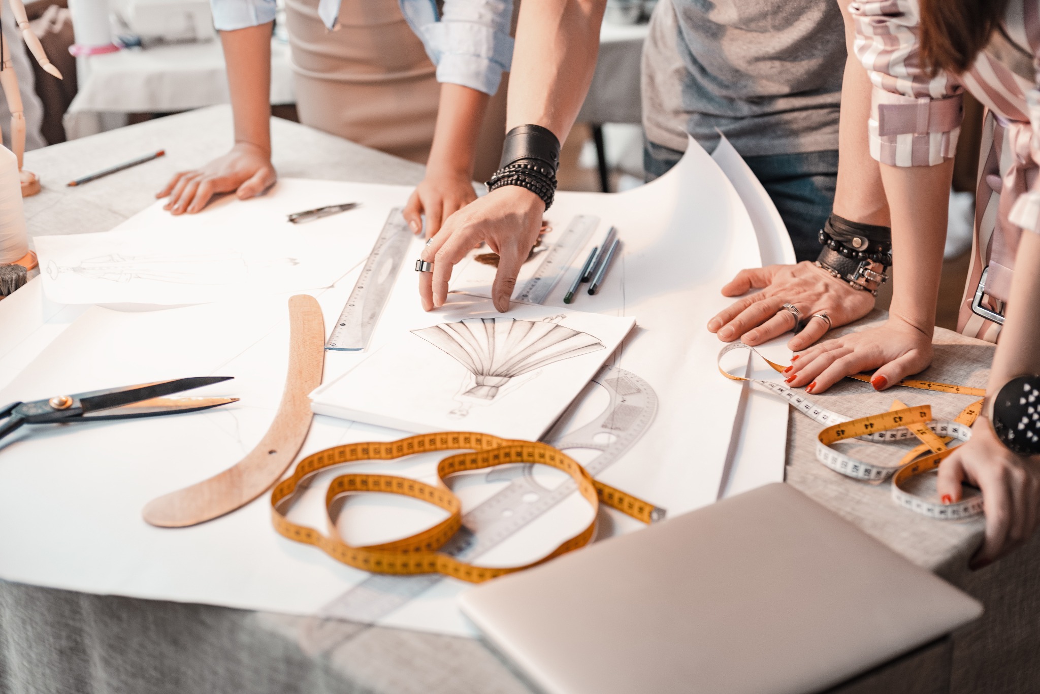 Zespół projektantów odzieży pracuje nad szkicami i pomiarami na stole z przyborami krawieckimi, widoczne detale: szkice sukienki, miara krawiecka, nożyczki i laptop.