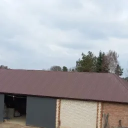 Budynek gospodarczy z nowym, brązowym dachem blaszanym, ceglano-białe ściany, szare, przesuwne wrota, widok z góry w pochmurny dzień.