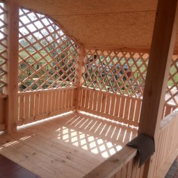 CHMUR-DACH Damian Chmura - Drewniana altana z kratownicą, widok z wnętrza, jasne drewno, wzór cienia na podłodze, fragment brązowej płytki na podłodze, widok na zewnątrz przez kratownicę.