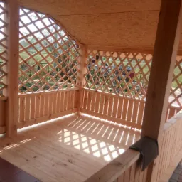 Drewniana altana z kratownicą, widok z wnętrza, jasne drewno, wzór cienia na podłodze, fragment brązowej płytki na podłodze, widok na zewnątrz przez kratownicę.