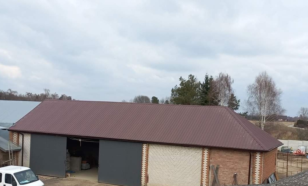 Budynek gospodarczy z nowym, brązowym dachem blaszanym, ceglano-białe ściany, szare, przesuwne wrota, widok z góry w pochmurny dzień.