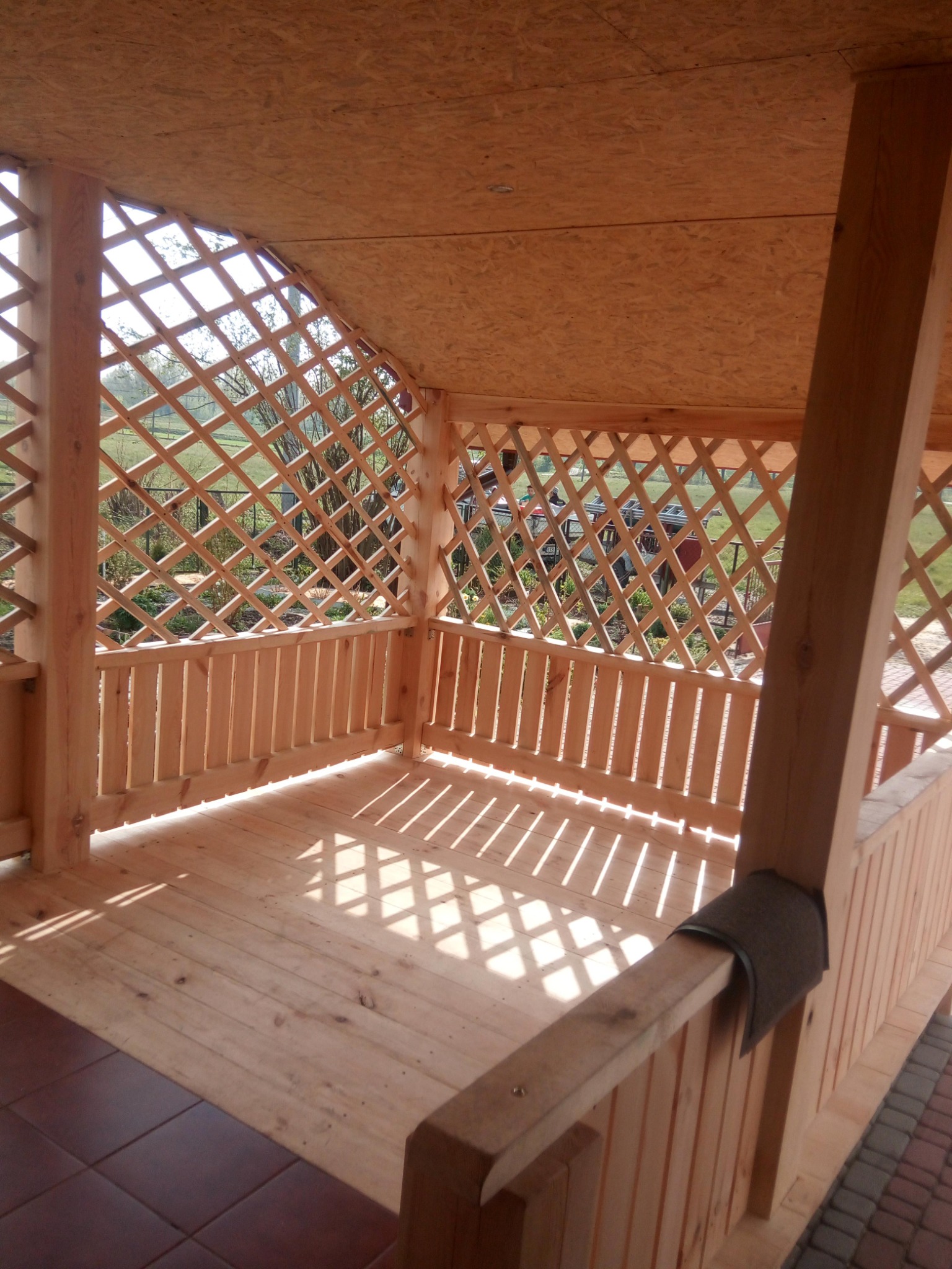 Drewniana altana z kratownicą, widok z wnętrza, jasne drewno, wzór cienia na podłodze, fragment brązowej płytki na podłodze, widok na zewnątrz przez kratownicę.