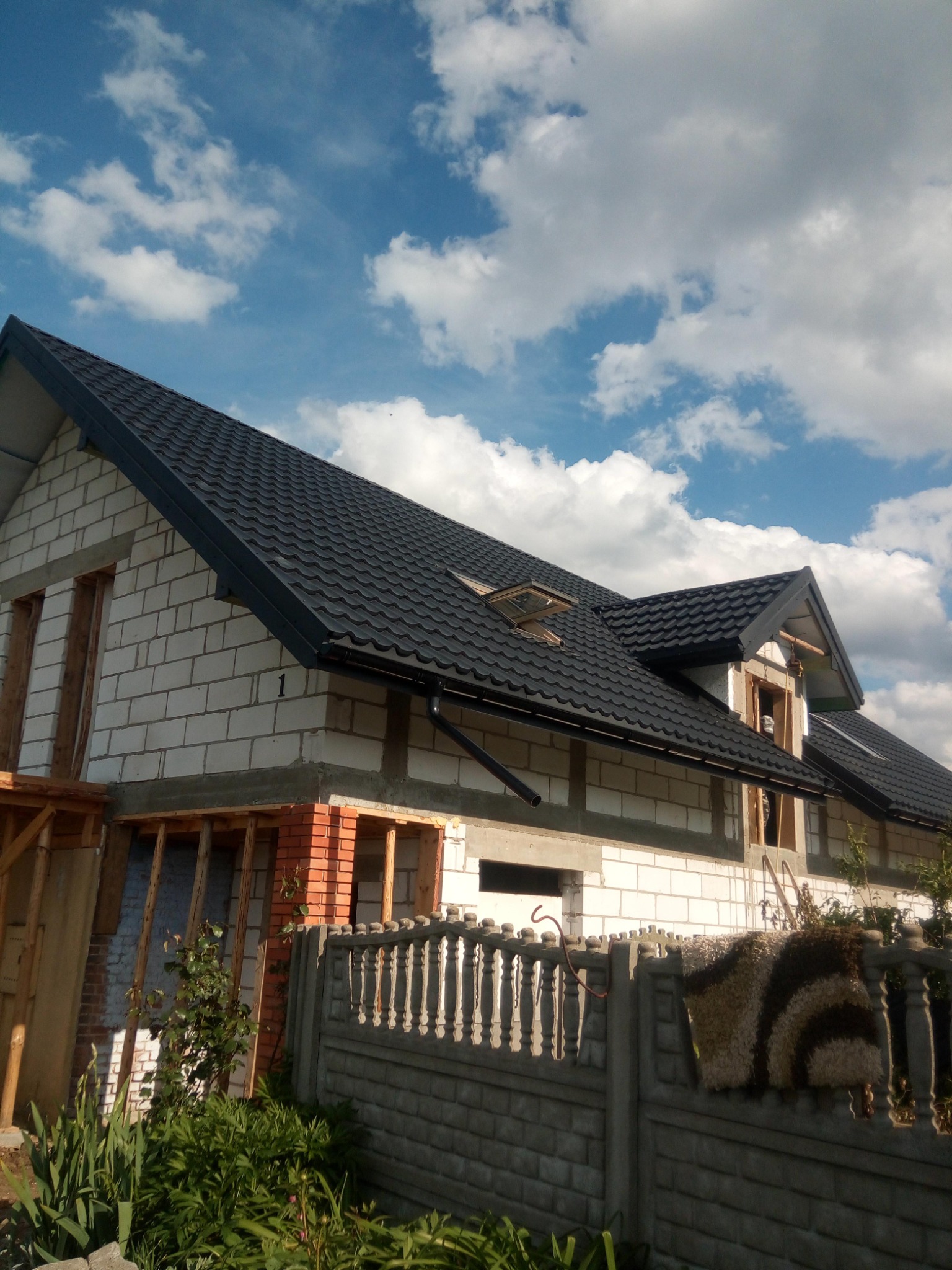 Dom w trakcie budowy z nowym, czarnym dachem z blachodachówki, widoczne okna dachowe i rynna, białe bloczki konstrukcyjne, czerwona cegła, ogrodzenie betonowe, błękitne niebo z chmurami.