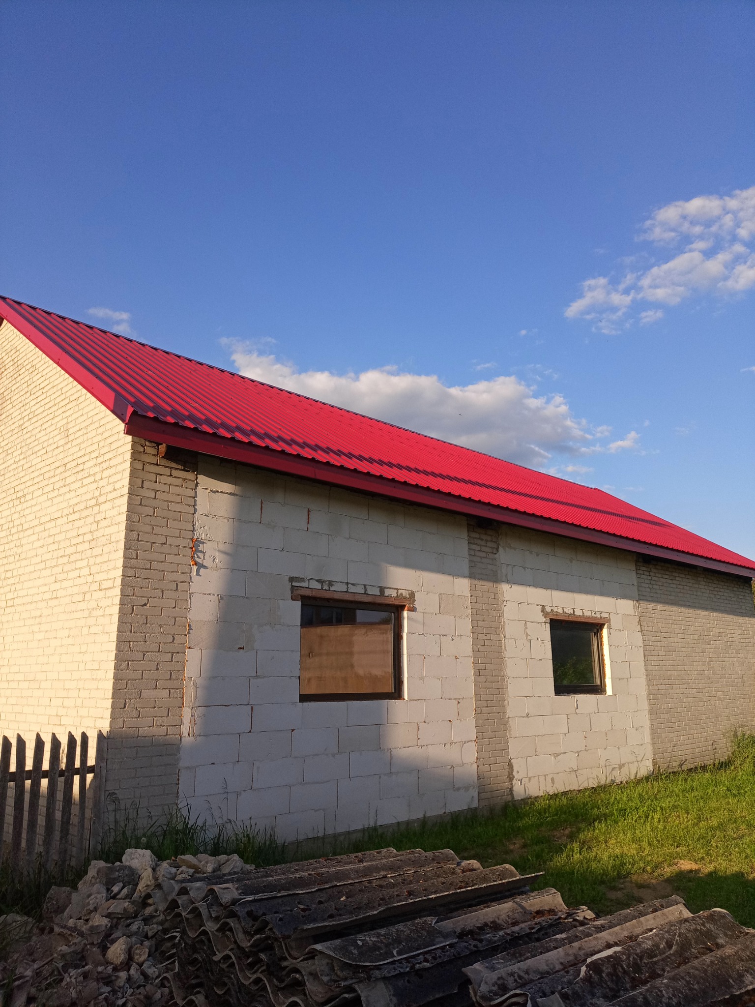 Świeżo położony, jaskrawoczerwony dach z blachy trapezowej na budynku z białych bloczków, częściowo otynkowanym, z widocznym stosem starych płyt eternitowych u podstawy ściany