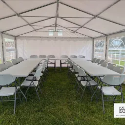 Wnętrze białego namiotu imprezowego z rozstawionymi białymi stołami i krzesłami na trawie, widoczne okna z przezroczystymi panelami.