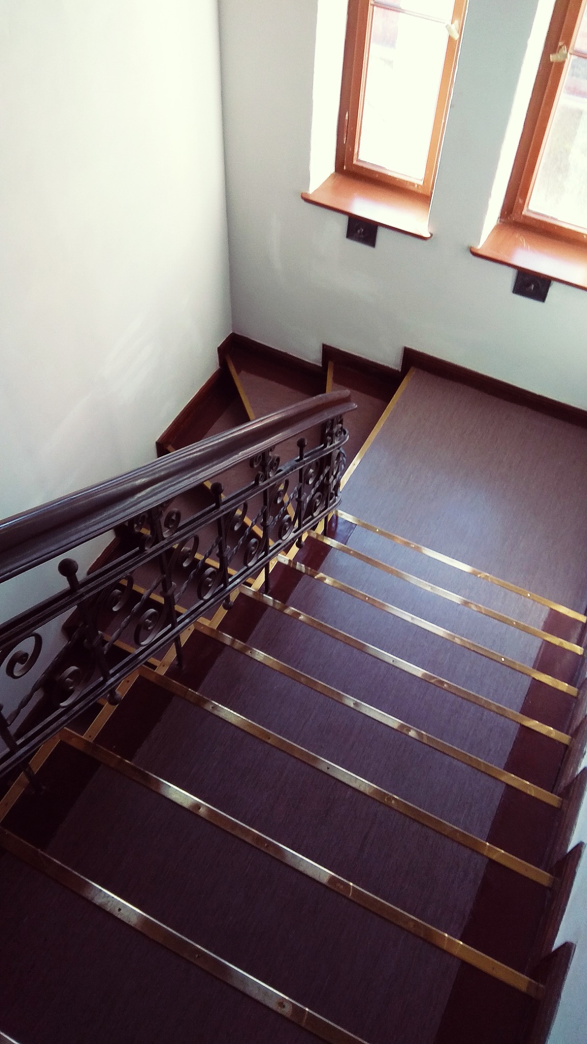 Wysoki kąt widzenia na odrestaurowane schody z ciemnego drewna z ozdobną metalową balustradą i złotymi listwami na stopniach, oświetlone przez dwa okna z drewnianymi ramami.