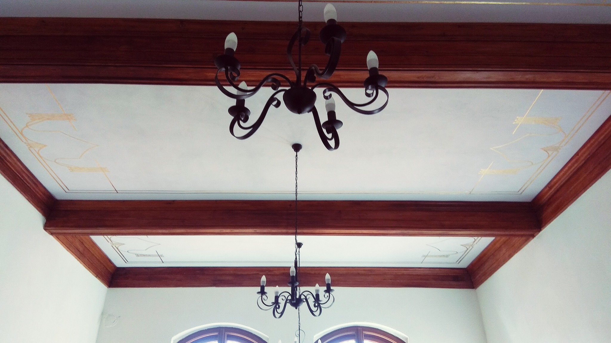 Odrestaurowany sufit z drewnianymi belkami i dekoracyjnymi, geometrycznymi malowidłami w kolorze złota, widoczne dwa żyrandole w stylu retro.