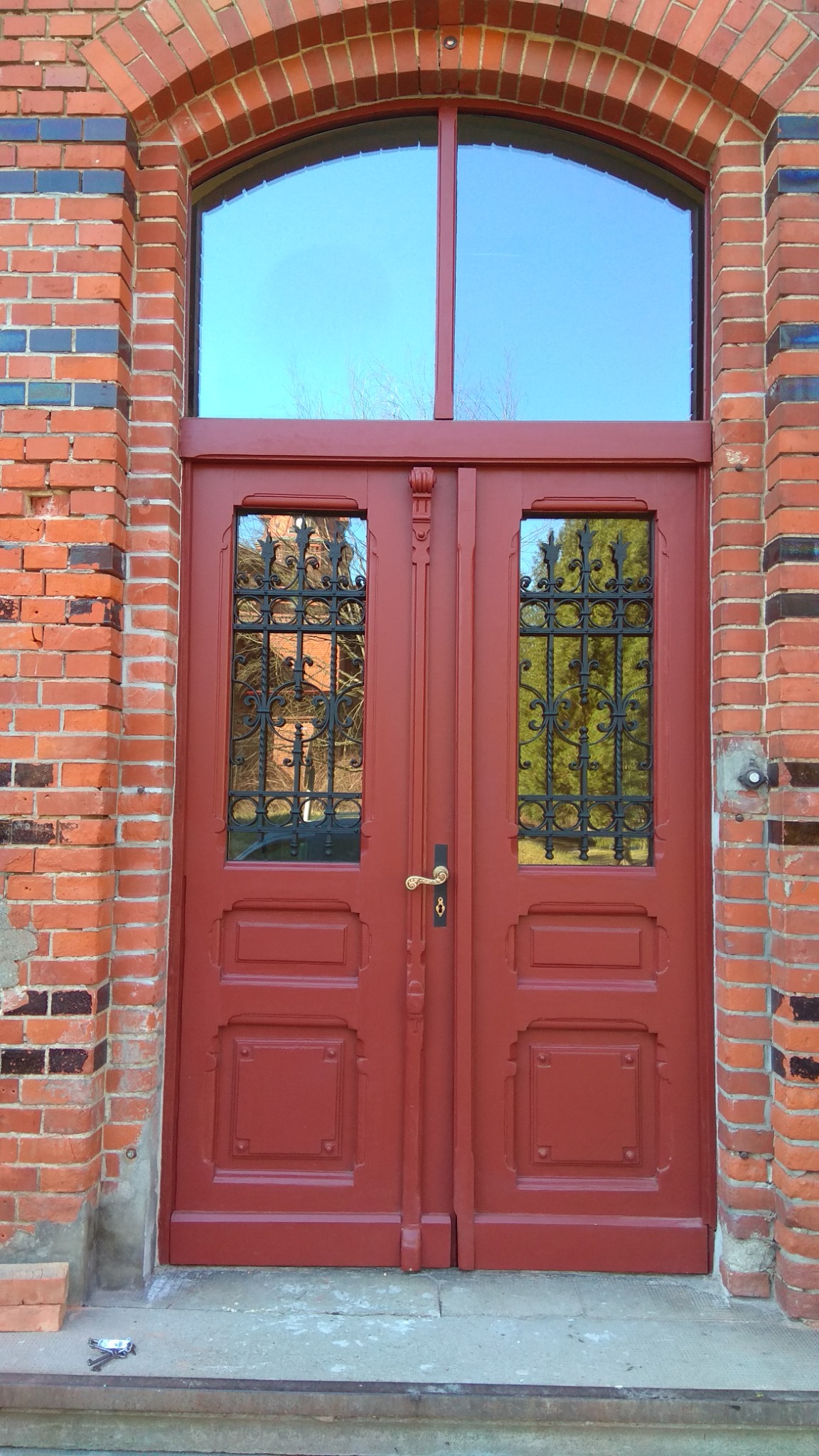 Odrestaurowane, czerwone, dwuskrzydłowe drzwi z ozdobnymi kratami w oknach, w Toruniu. Widoczny łuk ceglany nad drzwiami i fragment muru z różnokolorowej cegły.