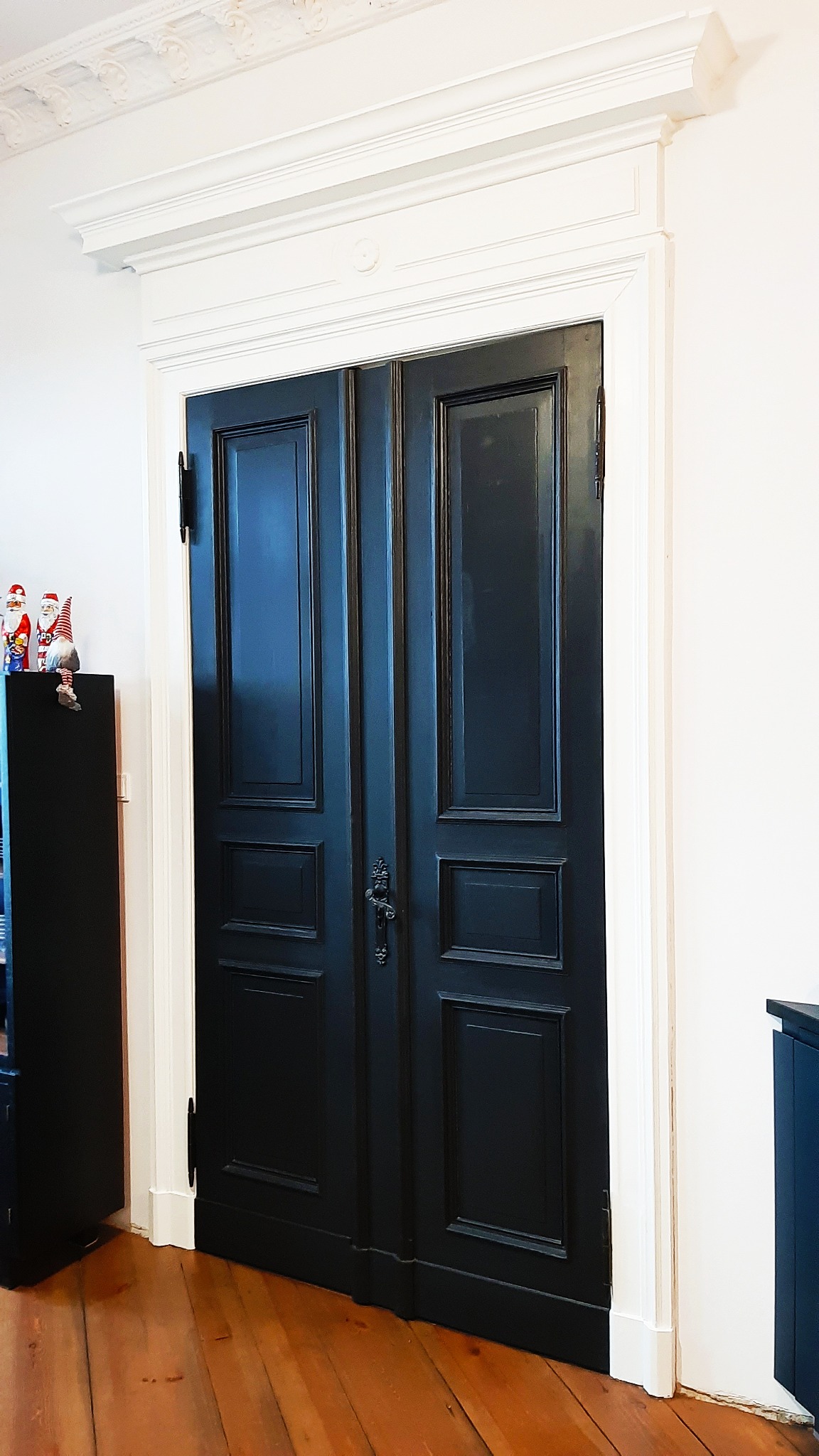 Odrestaurowane, dwuskrzydłowe, ciemnogranatowe drzwi z profilowanymi płycinami i ozdobnym zamkiem, osadzone w białej, klasycznej ościeżnicy z dekoracyjnym gzymsem, na tle drewnianej podłogi...