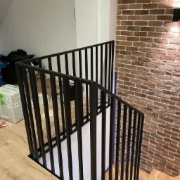 Nowoczesna, czarna balustrada metalowa na schodach z drewnianymi stopniami, obok ściana z cegły dekoracyjnej, widoczne narzędzia i materiały budowlane w tle.