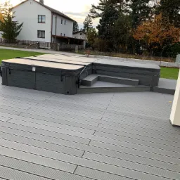 Dwa jacuzzi z pokrywami na szarym tarasie kompozytowym z dopasowanymi schodkami, widok na dom i drzewa w tle.