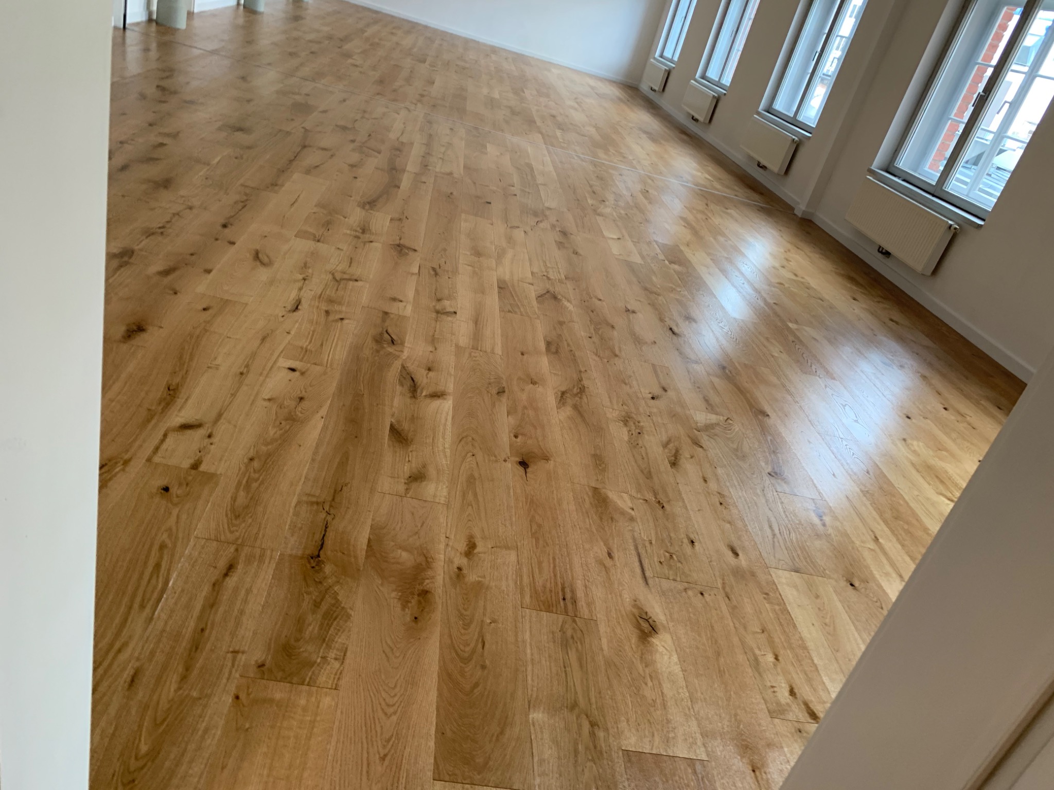 Jasna, drewniana podłoga z parkietu ułożona w przestronnym pokoju z białymi ścianami i kilkoma oknami, widok z perspektywy narożnika pokoju.