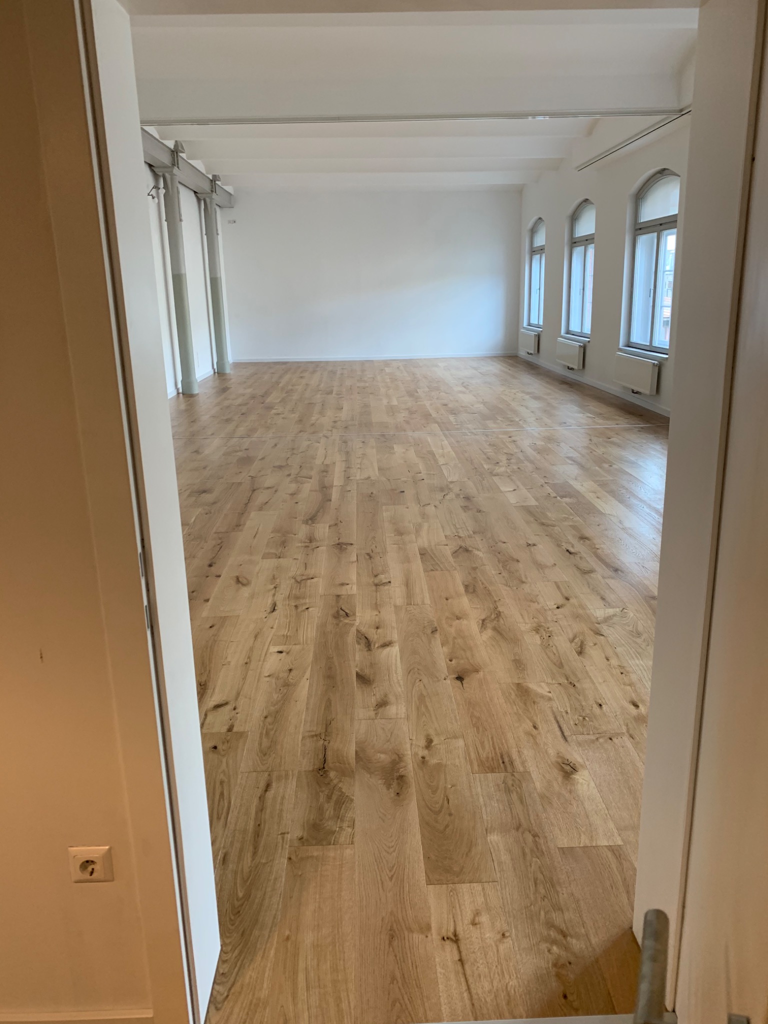 Jasne panele ułożone w przestronnym, minimalistycznym wnętrzu z białymi ścianami i wysokimi oknami łukowymi, widok zza otwartych drzwi.