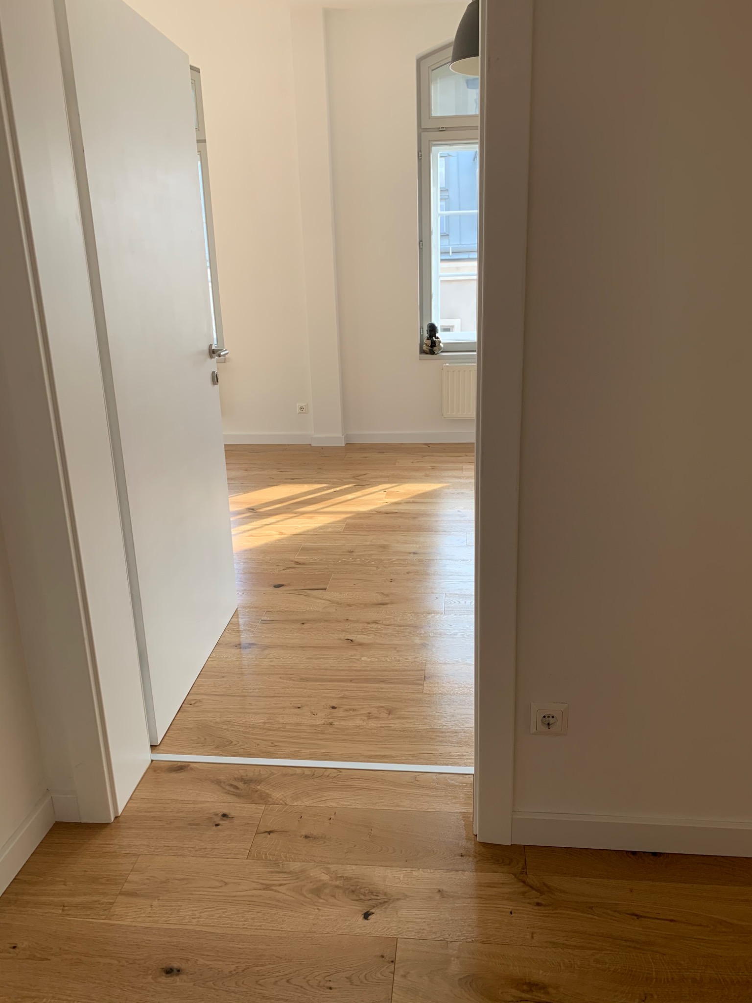 Jasne, dębowe panele podłogowe ułożone w nowym mieszkaniu, widoczne przez otwarte drzwi, z listwą przypodłogową i gniazdkiem elektrycznym przy ścianie.