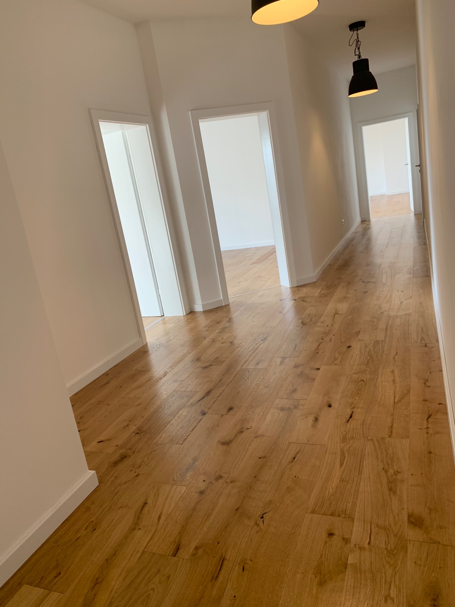 Długi korytarz z jasną, drewnianą podłogą ułożoną w jodełkę, białe ściany i drzwi, oświetlony nowoczesnymi czarnymi lampami wiszącymi.