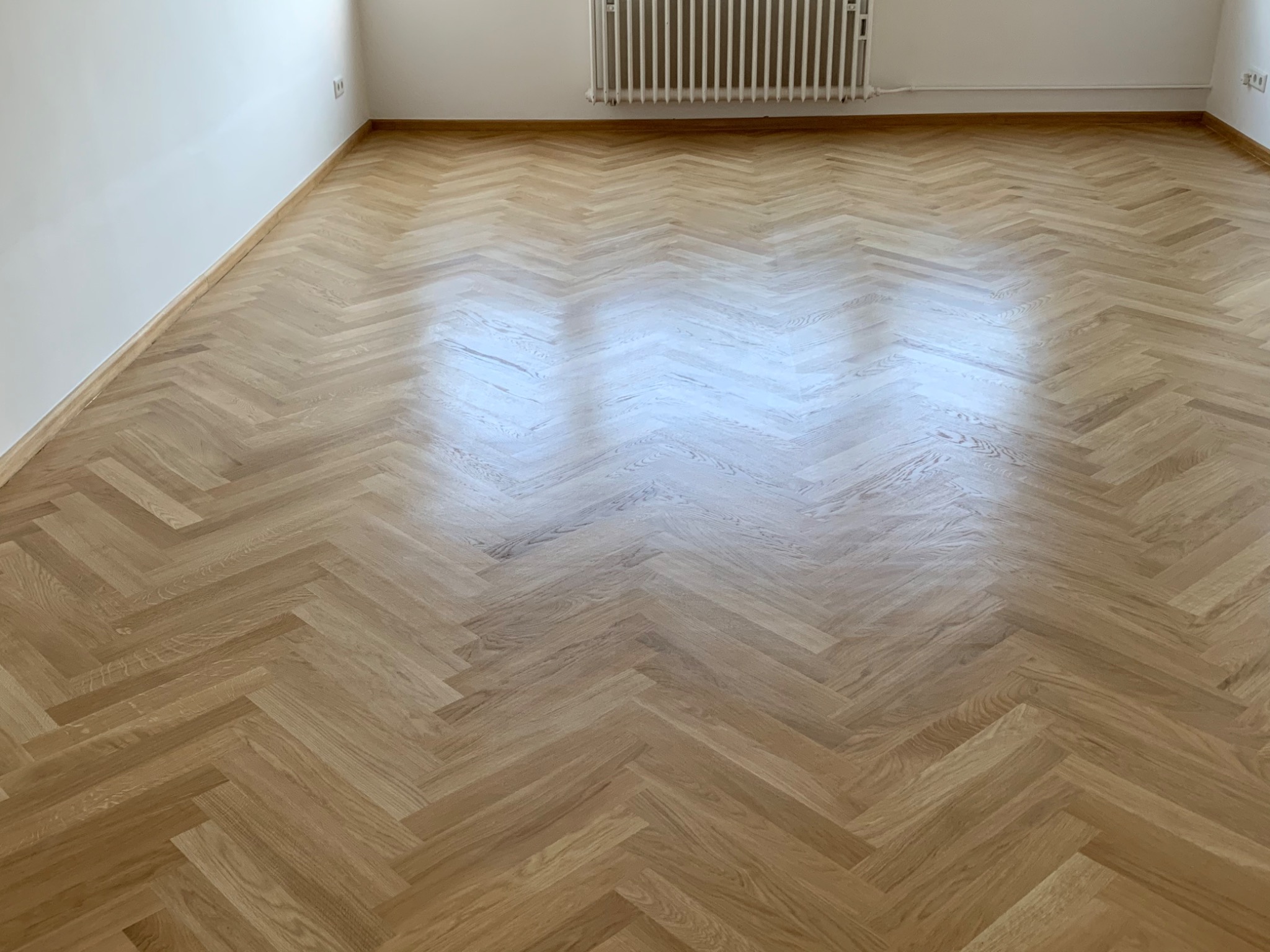 Jasny parkiet ułożony w jodełkę francuską, widoczny grzejnik na ścianie i listwy przypodłogowe.