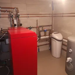 Instalacja centralnego ogrzewania z czerwonym kotłem, zbiornikiem na wodę i zmiękczaczem wody, rury miedziane zamocowane do ściany z płytek ceramicznych.