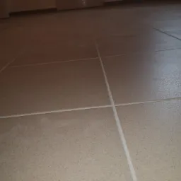 Beżowe płytki podłogowe z jasną fugą, ułożone równo, widoczne drzwi w tle.