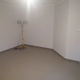 Pusty, świeżo wyremontowany pokój z szarą podłogą z płytek, oświetlony przenośną lampą budowlaną na żółtym statywie. Ściany pomalowane na jasny odcień szarości, widoczne gniazdko elektryczne. W tle...