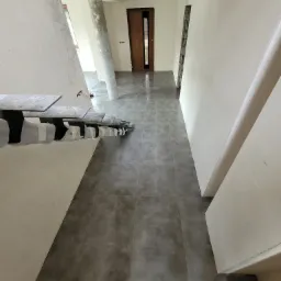 Korytarz w trakcie remontu z szarą, kwadratową podłogą z płytek, widoczne niedokończone schody z zabezpieczonymi stopniami i surowy betonowy filar.