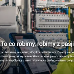 Elektryk w kasku i okularach ochronnych, analizujący schemat elektryczny na tablecie, z widoczną rozdzielnicą i okablowaniem w tle.