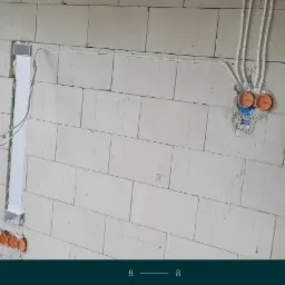 Wewnętrzna ściana z bloczków z widocznymi przewodami elektrycznymi w rurkach, puszkami instalacyjnymi i pionowym kanałem kablowym.