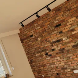 Ściana z czerwonej cegły z zamontowanym oświetleniem szynowym i wiszącą lampą z drewnianymi elementami, widoczne gniazdka elektryczne.