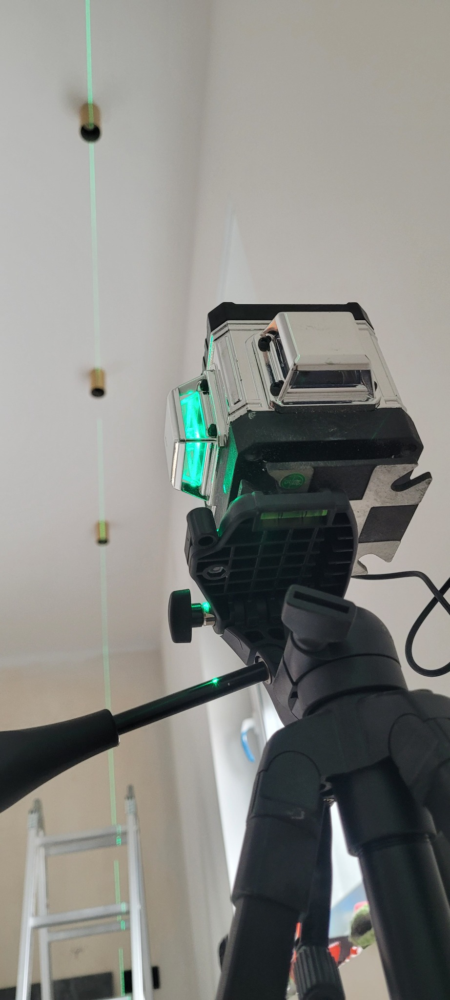 Laser krzyżowy na statywie, wyświetlający zielone linie na suficie z zamontowanymi puszkami pod oświetlenie, drabina w tle.