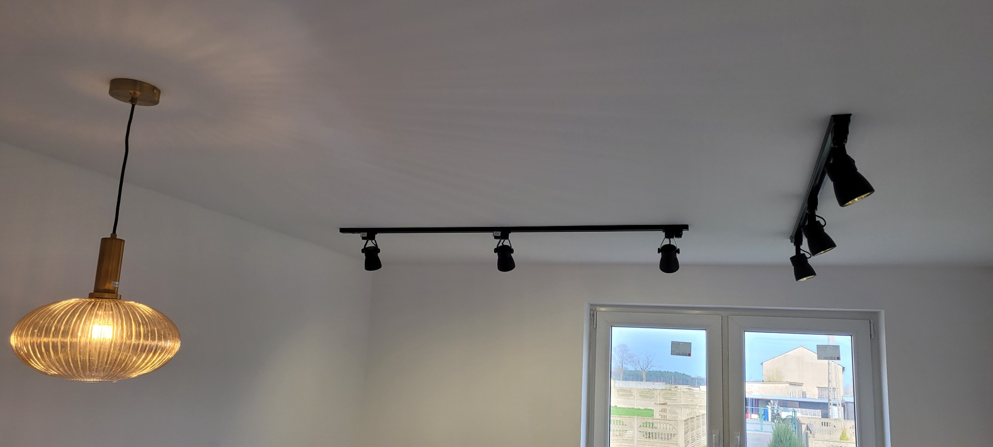 Nowoczesne oświetlenie sufitowe: złota lampa wisząca z żarówką filamentową oraz czarne reflektory szynowe w nowym, pomalowanym na biało pomieszczeniu z oknem.