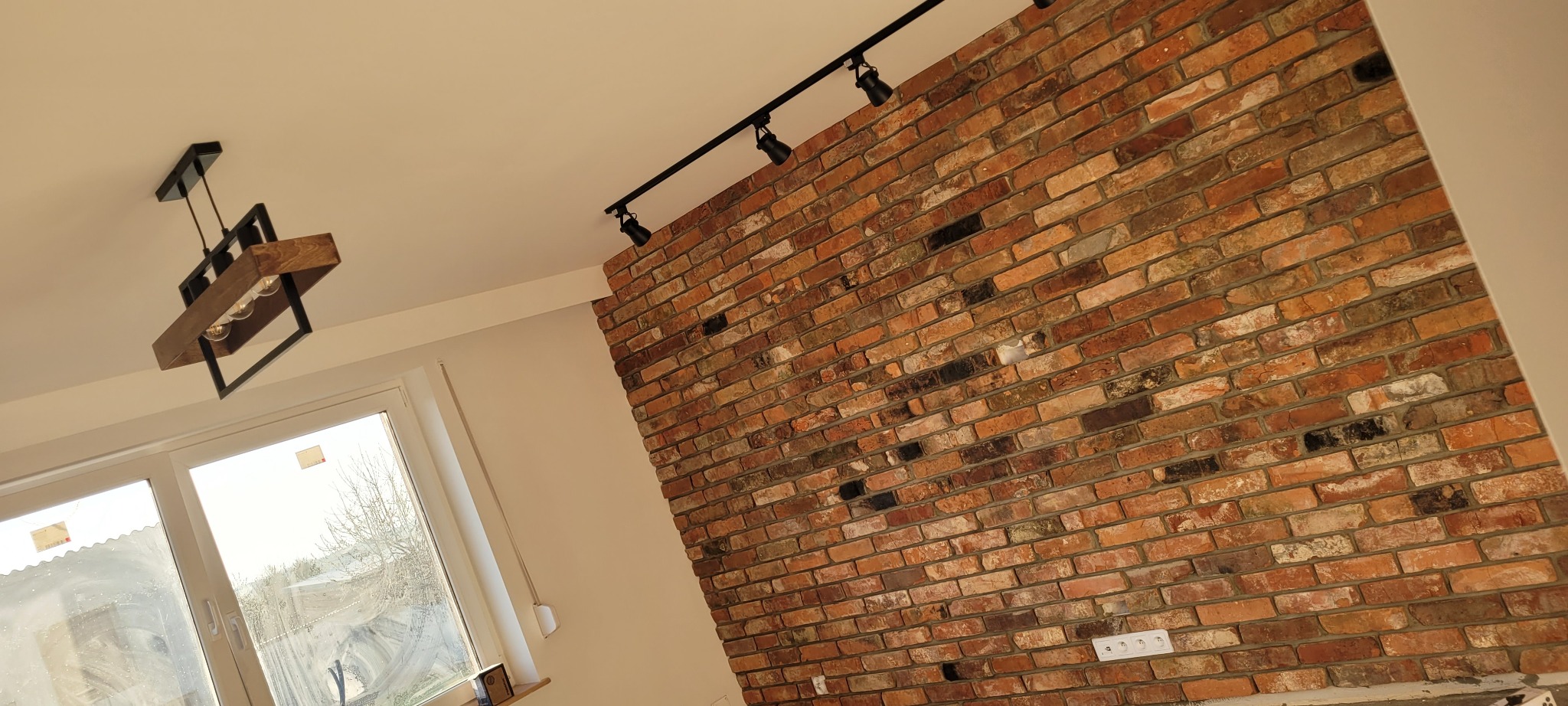 Ściana z czerwonej cegły z zamontowanym oświetleniem szynowym i wiszącą lampą z drewnianymi elementami, widoczne gniazdka elektryczne.