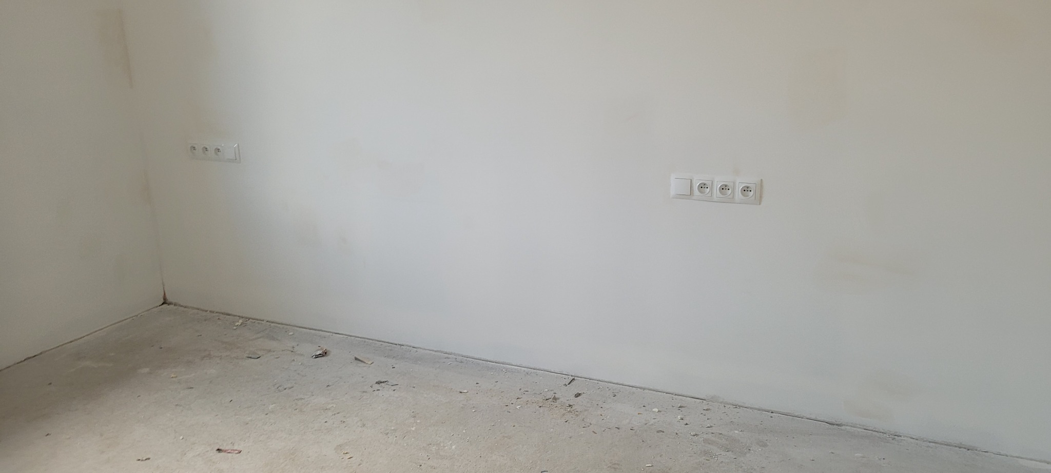 Surowe wnętrze z dwoma gniazdkami elektrycznymi i pojedynczym włącznikiem na świeżo pomalowanej ścianie, widoczny surowy beton na podłodze.