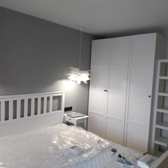 Pokój przed wykończeniem: białe meble (łóżko, szafa, szafka nocna) na tle szarej ściany, zabezpieczone folią malarską.