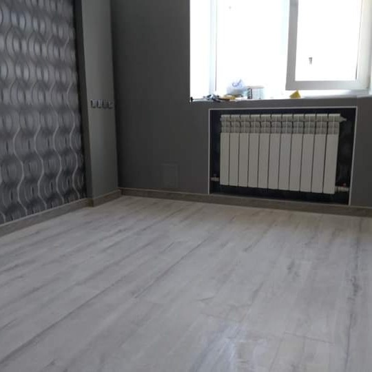 Pomalowane na szaro wnętrze pokoju z panelami podłogowymi w odcieniach bieli i szarości, grzejnik w zabudowie pod oknem, ściana z wzorzystą tapetą.