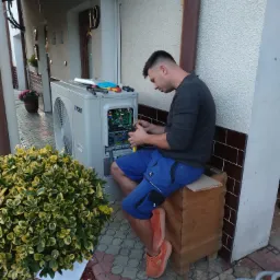 Instalator w niebieskich spodenkach siedzi na drewnianej skrzynce i podłącza kable w otwartej obudowie białej pompy ciepła York na zewnątrz budynku. Obok leżą narzędzia i zwinięty szary przewód.