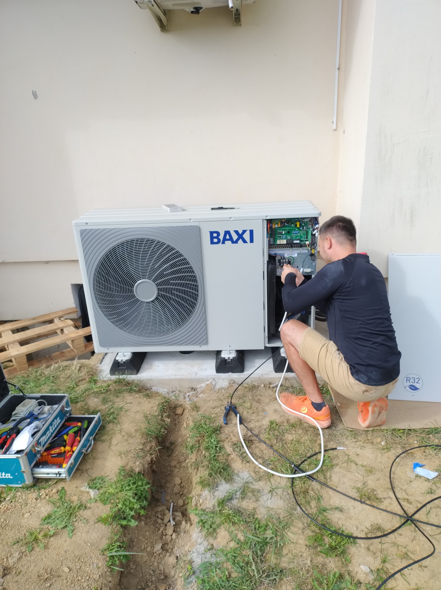 Instalator w pomarańczowych butach podłącza kable w otwartej obudowie pieca gazowego Baxi, skrzynka narzędziowa Makita obok wykopu.
