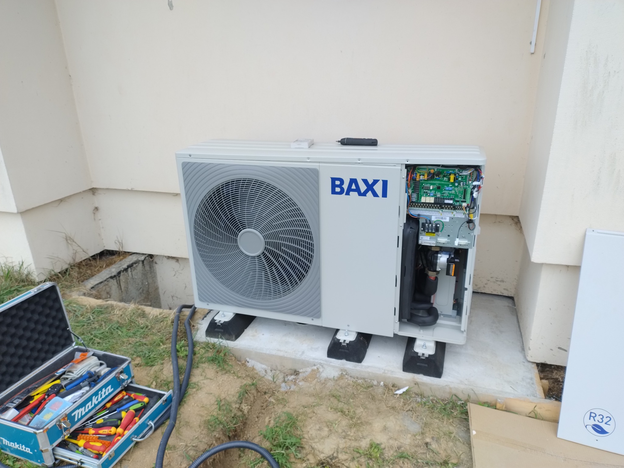 Biała pompa ciepła marki BAXI, z otwartą obudową, ukazującą wewnętrzne komponenty elektroniczne, stoi na betonowej podstawie obok skrzynki narzędziowej Makita z widocznymi śrubokrętami i szczypcami.