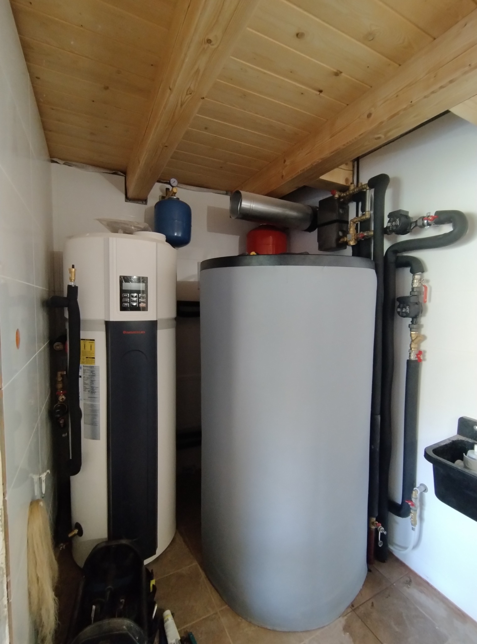 Dwa zbiorniki na wodę w pomieszczeniu z drewnianym sufitem, rury przyłączeniowe, instalacja wodna, widoczny manometr na zbiorniku.