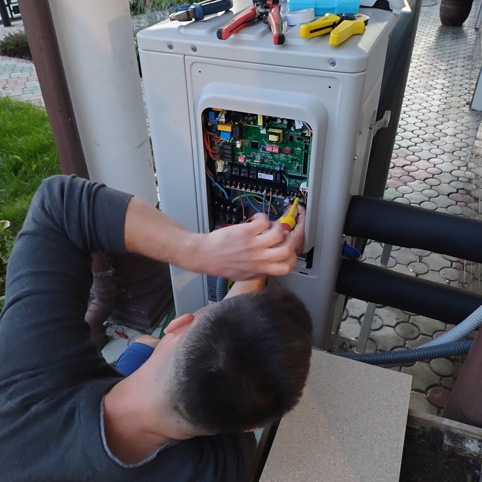 Instalator w trakcie podłączania przewodów wewnątrz obudowy jednostki zewnętrznej klimatyzacji lub pompy ciepła, widoczne narzędzia na górnej powierzchni urządzenia.