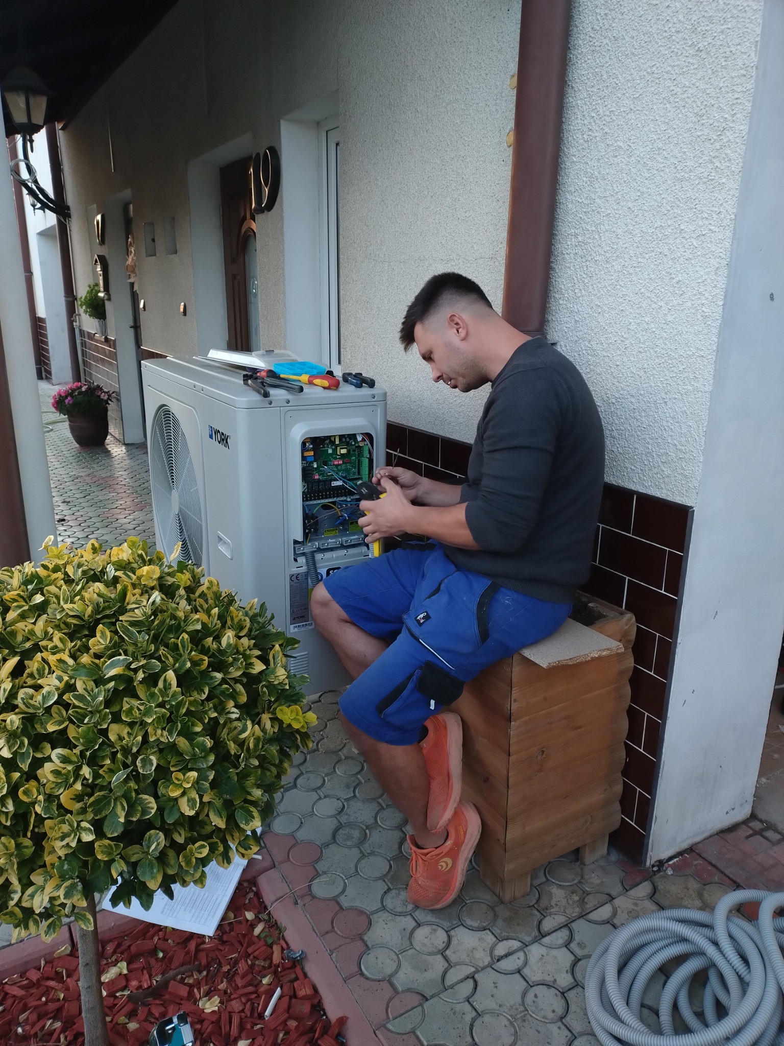 Instalator w niebieskich spodenkach siedzi na drewnianej skrzynce i podłącza kable w otwartej obudowie białej pompy ciepła York na zewnątrz budynku. Obok leżą narzędzia i zwinięty szary przewód.