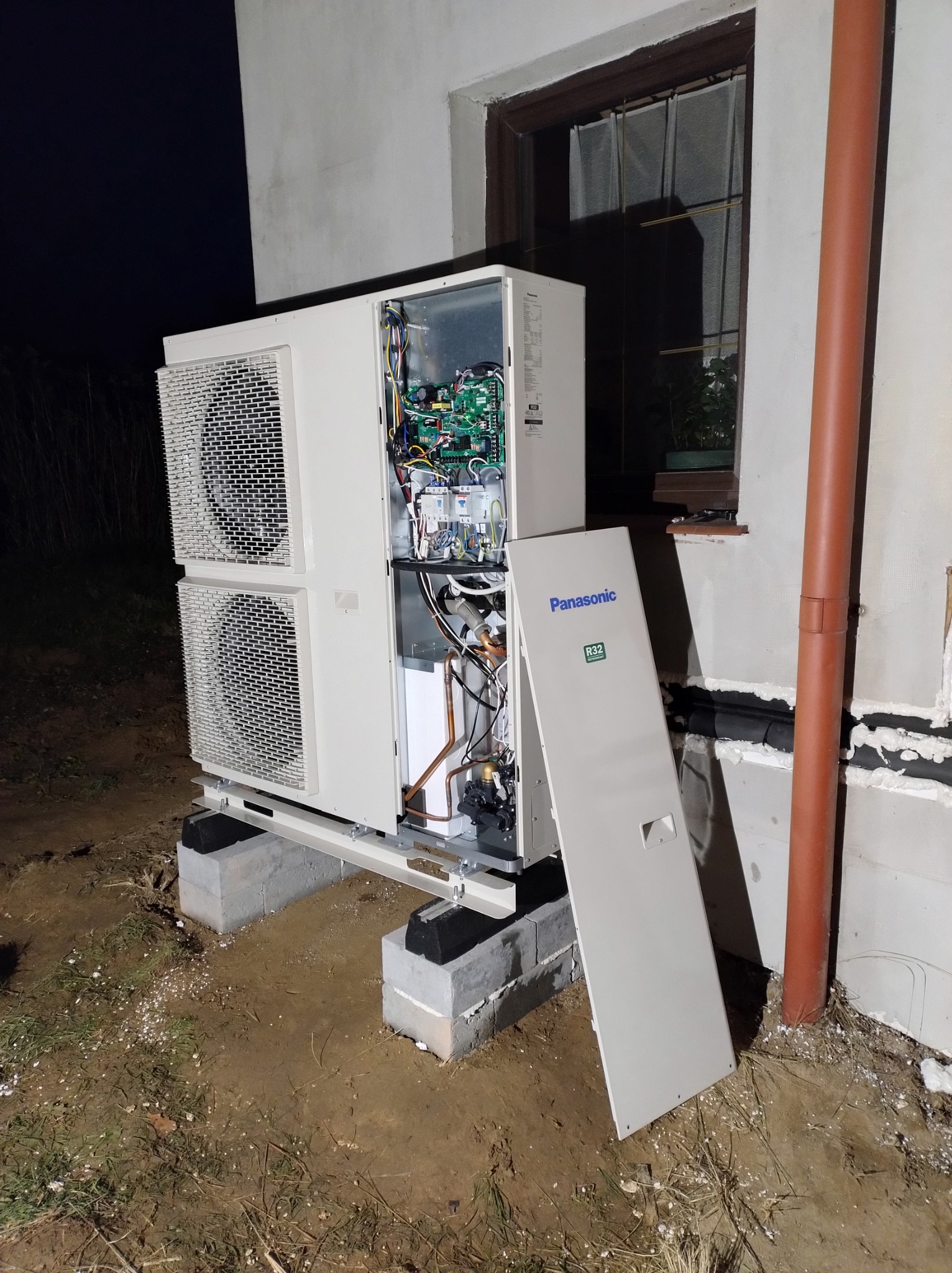 Zewnętrzna jednostka pompy ciepła Panasonic R32 na betonowych bloczkach, z otwartą obudową ukazującą wewnętrzne komponenty, widoczna instalacja przy ścianie budynku.