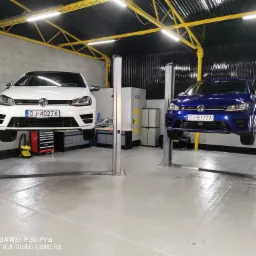 Dwa samochody VW Golf R, biały i niebieski, podniesione na podnośnikach w jasnym warsztacie samochodowym z żółtą konstrukcją sufitu i czarnymi ścianami w tle.