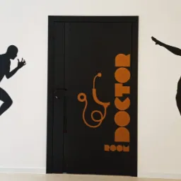 Ściana w gabinecie lekarskim z czarnymi sylwetkami biegacza i siatkarza, drzwi z napisem 'Doctor Room' i symbolem stetoskopu w kolorze pomarańczowym.