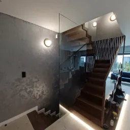 Schody drewniane z szklaną balustradą w nowoczesnym domu w Garwolinie, ściana wykończona dekoracyjną farbą imitującą beton.