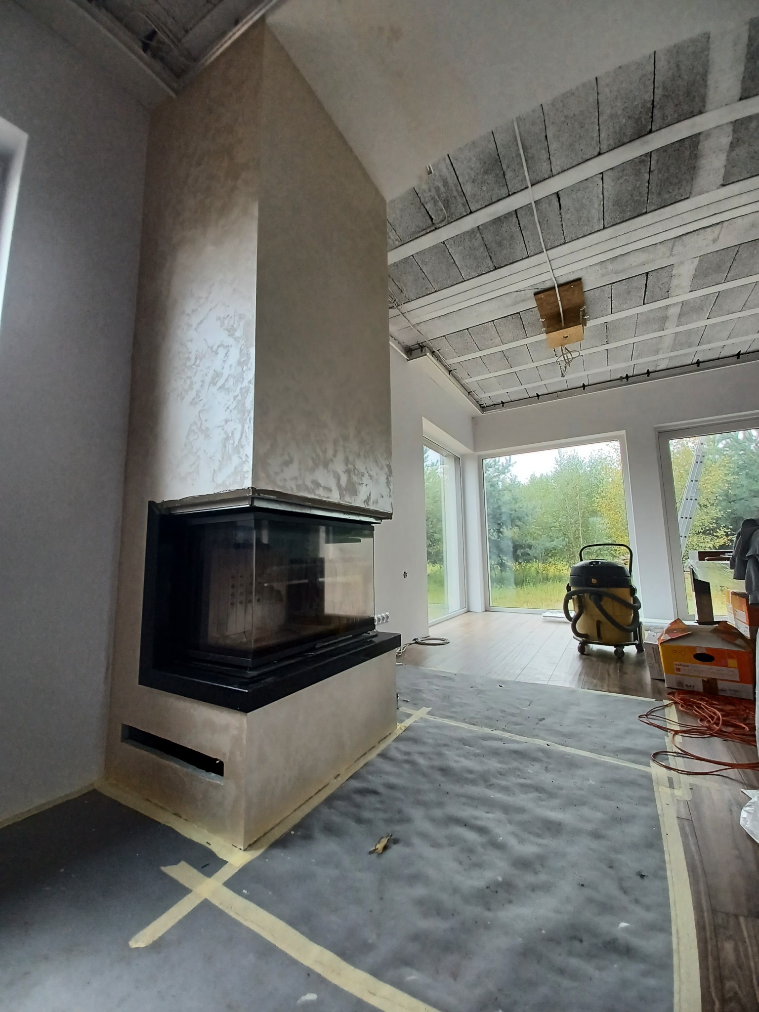 Wykończony kominek z obudową z dekoracyjną fakturą, zabezpieczona podłoga, widoczny odkurzacz przemysłowy i surowy sufit z instalacjami.