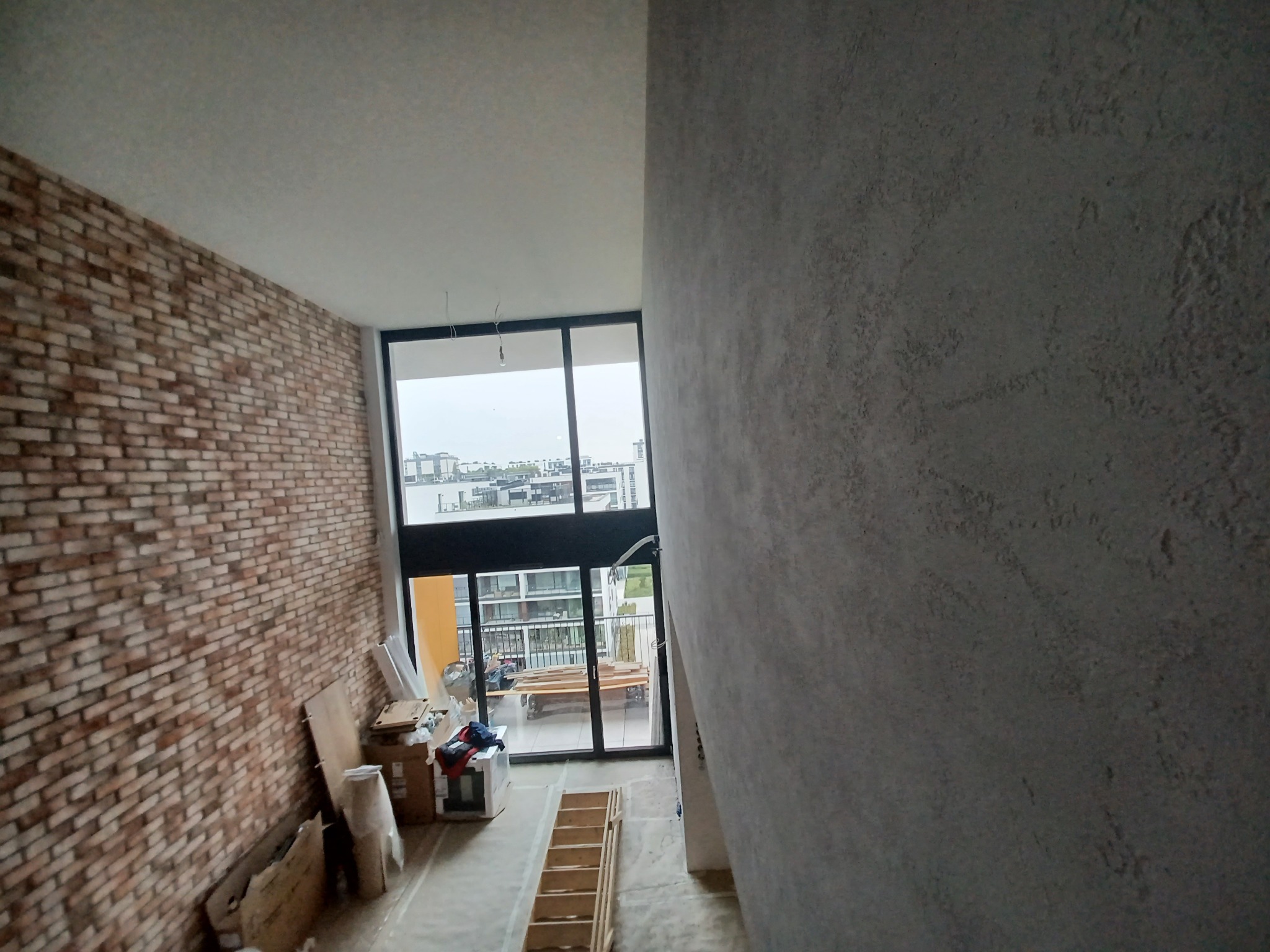 Surowa ściana z efektem betonu w jasnoszarym kolorze, kontrastująca z ceglaną ścianą i dużym oknem balkonowym w tle, drabina i kartony na podłodze.