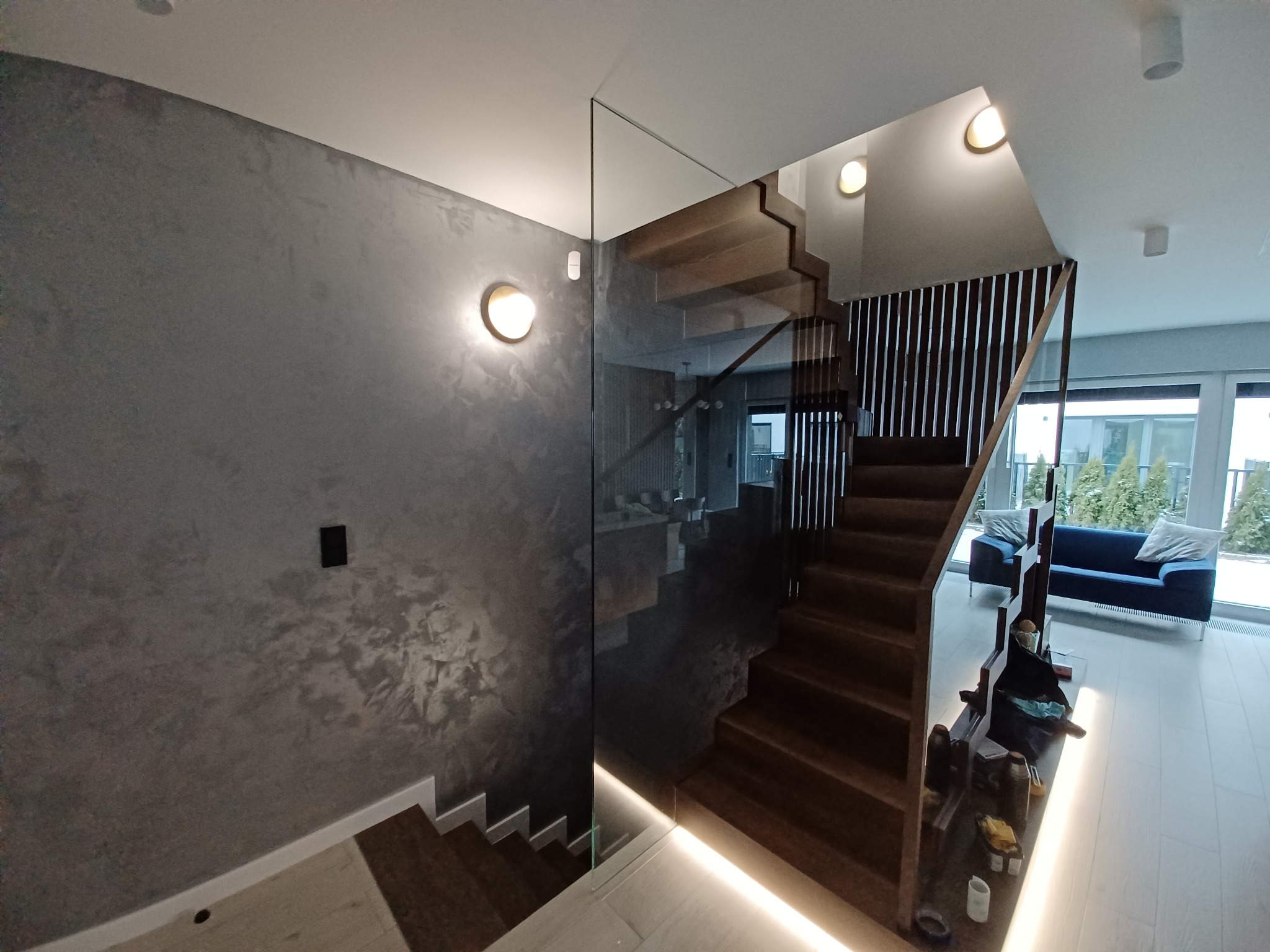 Schody drewniane z szklaną balustradą w nowoczesnym domu w Garwolinie, ściana wykończona dekoracyjną farbą imitującą beton.