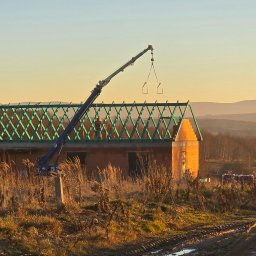 SZYMI-BUD - Wznoszenie domu z cegły: dźwig podnosi element konstrukcyjny na zieloną więźbę dachową. W tle pagórkowaty krajobraz w ciepłym świetle zachodzącego słońca.