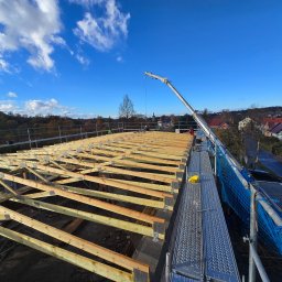 SZYMI-BUD - Wznoszenie drewnianej konstrukcji dachu z użyciem dźwigu na tle błękitnego nieba. Widoczne rusztowania i elementy zabezpieczeń.