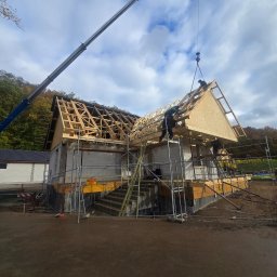 SZYMI-BUD - Montaż prefabrykowanej więźby dachowej na budynku z bloczków, z użyciem dźwigu. Widoczni pracownicy na rusztowaniach. Tło: drzewa i pochmurne niebo.