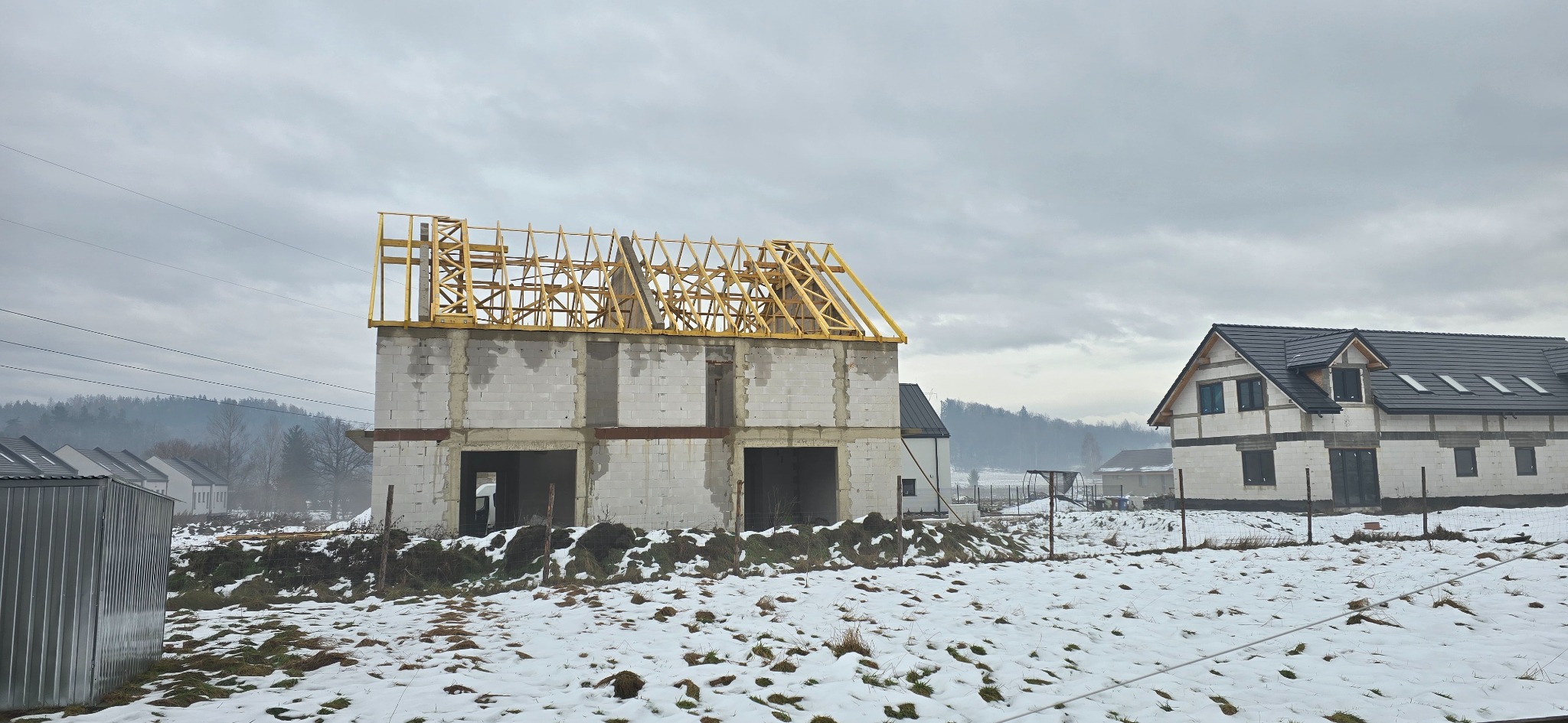 Budowa domu w zimowej scenerii: surowy stan z widoczną drewnianą konstrukcją dachu, obok dom z ciemnym dachem. W tle zalesione wzgórza pod pochmurnym niebem.